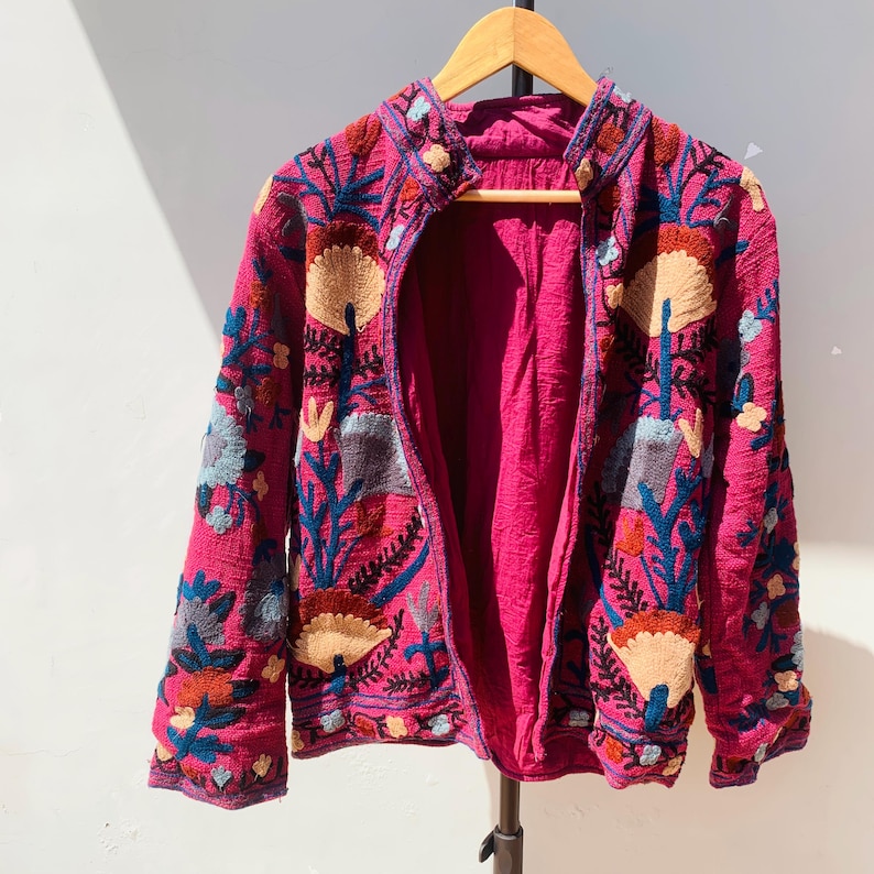 Op de afbeelding: Een magenta jasje met ingewikkelde bloemenborduurwerk in blauw, beige en rood. Het open jasje onthult een magenta voering. Het borduurwerk toont gestileerde bloemen en bladeren, wat een bohemien esthetiek cre&euml;ert. Het jasje hangt aan een houten hanger.