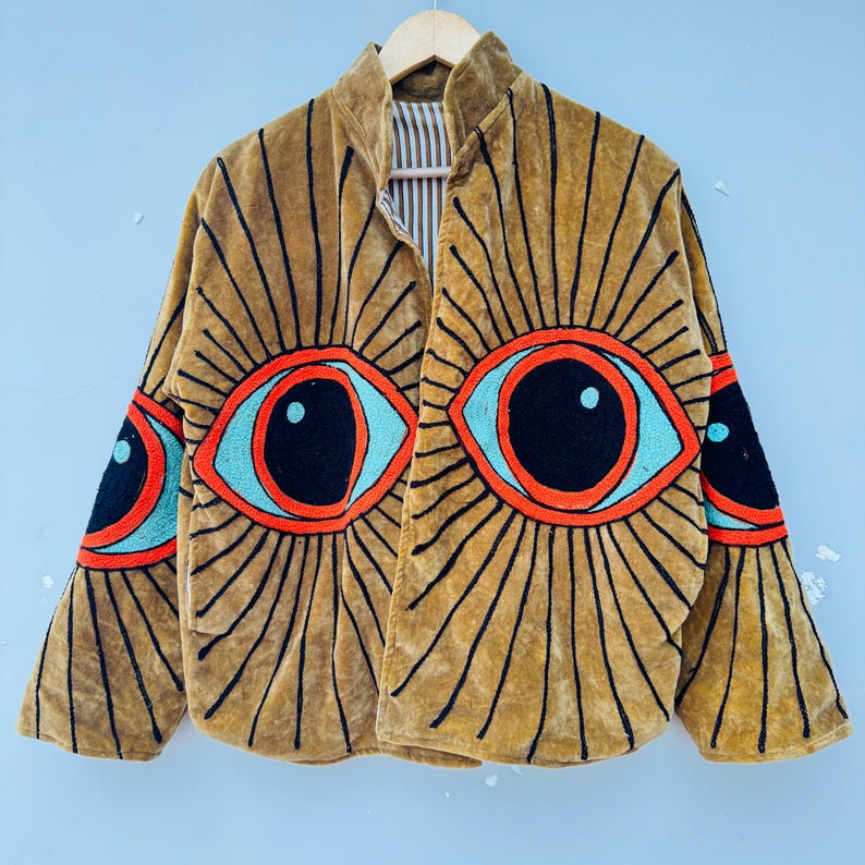 May include: Mustard-colored velvet jacket with embroidered eye designs. The jacket features large, stylized eyes in red, black, and turquoise, radiating black lines. The jacket has a high collar and is hanging on a wooden hanger.