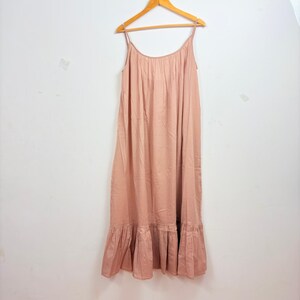 May include: A light pink maxi dress with spaghetti straps, hanging on a wooden hanger. The dress has a gathered bodice and a ruffled hemline. The fabric appears to be lightweight and flowing, suitable for warm weather.