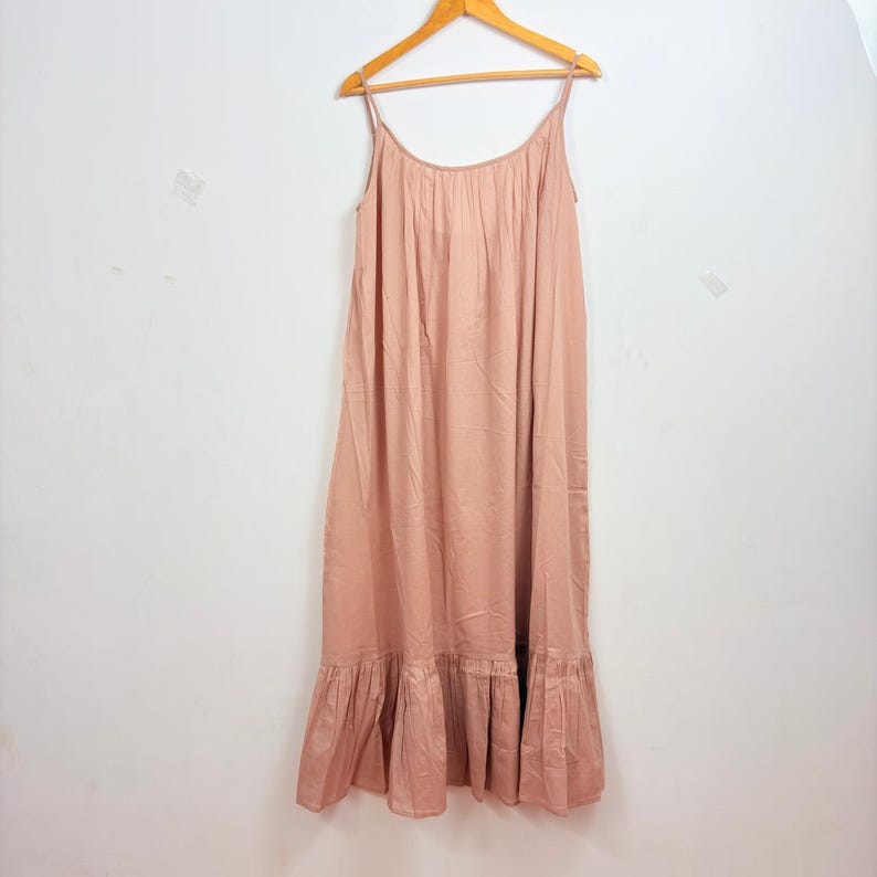 May include: A light pink maxi dress with spaghetti straps, hanging on a wooden hanger. The dress has a gathered bodice and a ruffled hemline. The fabric appears to be lightweight and flowing, suitable for warm weather.