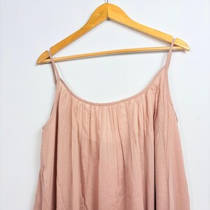 May include: A light pink camisole top with thin straps, hanging on a wooden hanger. The top has a gathered neckline and a flowing, loose fit. The fabric appears to be a soft, lightweight material.