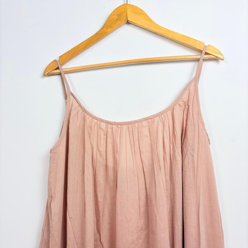 May include: A light pink camisole top with thin straps, hanging on a wooden hanger. The top has a gathered neckline and a flowing, loose fit. The fabric appears to be a soft, lightweight material.