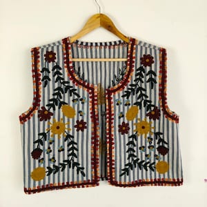 May include: A striped vest with floral embroidery. The vest has vertical blue and white stripes, with embroidered flowers in yellow, burgundy, and blue. The edges are trimmed with a red and orange pom-pom trim.