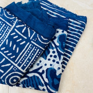 May include: Indigo blue fabric with white geometric patterns and stripes. The fabric is folded and stacked, showcasing various designs. The patterns include lines, dots, and abstract shapes, creating a visually appealing textile.