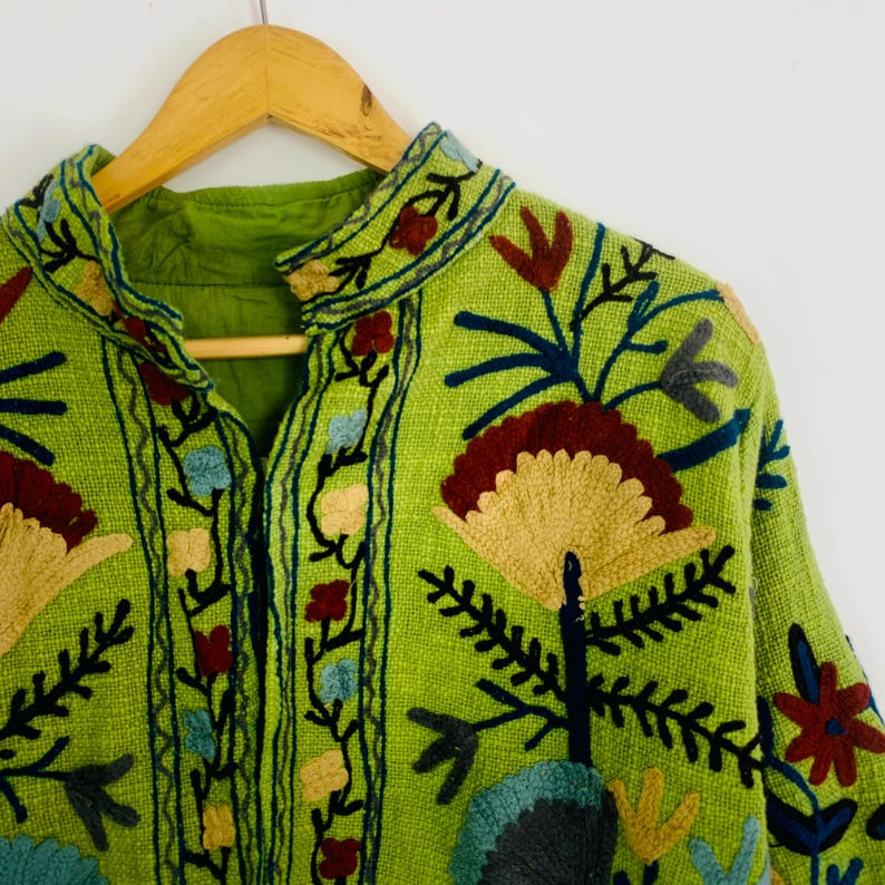 May include: A lime green jacket with intricate floral embroidery in shades of red, yellow, blue, and gray. The jacket features a mandarin collar and is hanging on a wooden hanger. The embroidery creates a textured, artistic design.