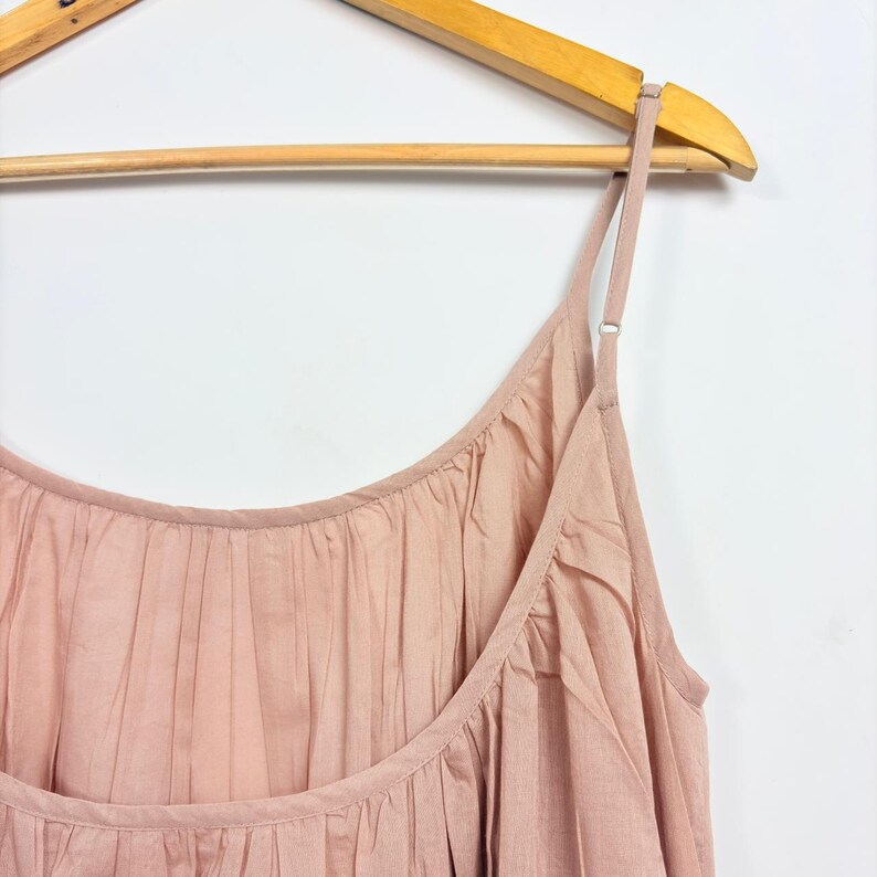 May include: A light pink camisole dress with adjustable spaghetti straps hangs on a wooden hanger. The dress features a gathered neckline and a flowing, pleated design. The fabric appears to be lightweight and suitable for warm weather.