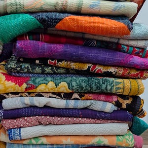 Könnte beinhalten: Ein Stapel bunter, gemusterter Quilts. Die Quilts zeigen eine Vielzahl von Farben, darunter Grün, Orange, Lila und Blau. Jeder Quilt hat ein einzigartiges Design mit sichtbaren Nähten und Mustern. Die Quilts sind ordentlich gestapelt und zeigen ihre Texturen und Designs.