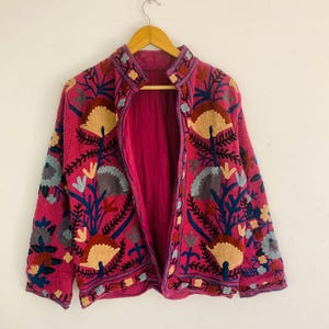 Op de afbeelding: Een magenta jasje met gedetailleerde bloemenborduursels in beige, blauw en groen. De jas heeft een opstaande kraag en is volledig gevoerd met magenta stof. De borduursels zijn ingewikkeld en bedekken de voorkant, mouwen en kraag.