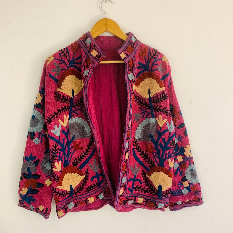 Op de afbeelding: Een magenta jasje met gedetailleerde bloemenborduursels in beige, blauw en groen. De jas heeft een opstaande kraag en is volledig gevoerd met magenta stof. De borduursels zijn ingewikkeld en bedekken de voorkant, mouwen en kraag.
