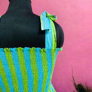 Puede incluir: Vestido sin mangas con estampado de rayas azules y verdes. El vestido tiene tirantes anudados y un escote con volantes. La tela est&aacute; fruncida, creando un efecto texturizado. El vestido se exhibe en un maniqu&iacute; negro sobre un fondo rosa.