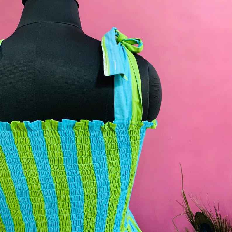 Puede incluir: Vestido sin mangas con estampado de rayas azules y verdes. El vestido tiene tirantes anudados y un escote con volantes. La tela est&aacute; fruncida, creando un efecto texturizado. El vestido se exhibe en un maniqu&iacute; negro sobre un fondo rosa.