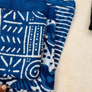 May include: Folded indigo blue fabric with white geometric and striped patterns. The fabric appears to be cotton or a similar natural fiber. The designs include various shapes and lines, creating a visually interesting textile.