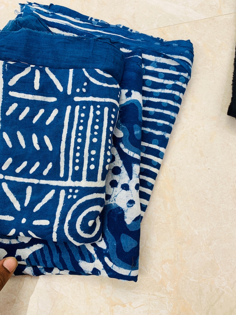 May include: Folded indigo blue fabric with white geometric and striped patterns. The fabric appears to be cotton or a similar natural fiber. The designs include various shapes and lines, creating a visually interesting textile.