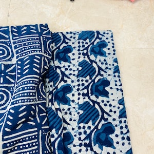 May include: Two pieces of fabric with indigo blue and white patterns. One features geometric shapes and lines, the other has a floral design with leaves and dots. The fabrics are folded and lying on a light-colored surface.