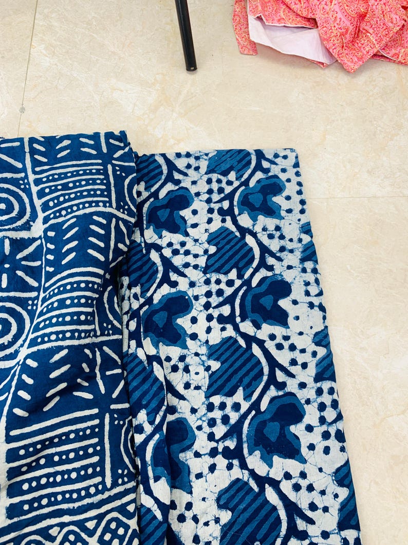 May include: Two pieces of fabric with indigo blue and white patterns. One features geometric shapes and lines, the other has a floral design with leaves and dots. The fabrics are folded and lying on a light-colored surface.