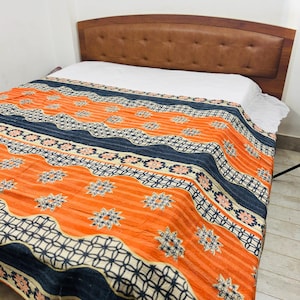 Può includere: Un letto con testiera in pelle marrone e una coperta fantasia arancione, blu navy e bianca. La coperta presenta strisce e motivi floreali. Il letto è coperto da un lenzuolo bianco.