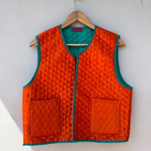 Puede incluir: Un chaleco acolchado naranja con forro y ribete turquesa, colgado de una percha de madera. El chaleco tiene dos bolsillos delanteros y un patrón de diamantes. Prenda sin mangas.