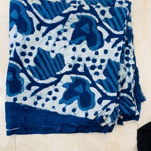 May include: A folded textile with a blue and white pattern. The design features organic shapes, dots, and stripes in varying shades of blue against a white background. The fabric is folded, revealing layers of the patterned material.