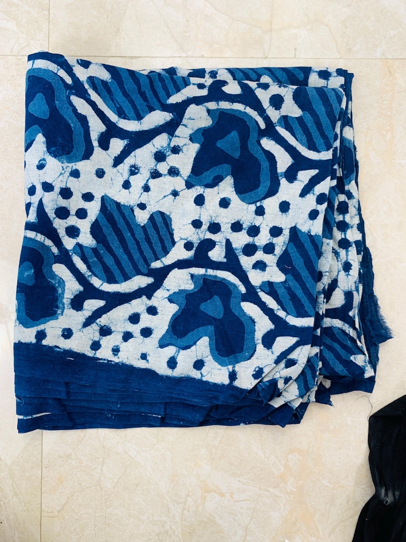 May include: A folded textile with a blue and white pattern. The design features organic shapes, dots, and stripes in varying shades of blue against a white background. The fabric is folded, revealing layers of the patterned material.