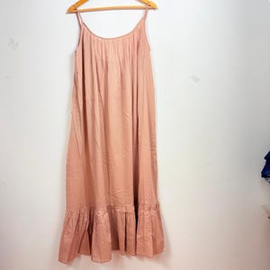 May include: A light pink maxi dress with spaghetti straps, hanging on a wooden hanger. The dress has a gathered bodice and a ruffled hemline. The fabric appears to be lightweight and flowing, suitable for warm weather.