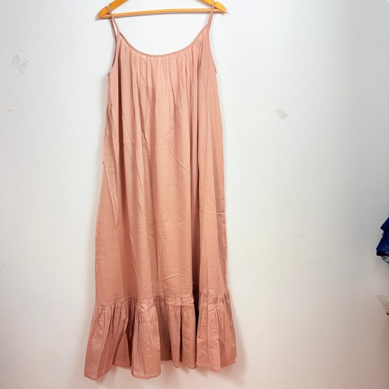 May include: A light pink maxi dress with spaghetti straps, hanging on a wooden hanger. The dress has a gathered bodice and a ruffled hemline. The fabric appears to be lightweight and flowing, suitable for warm weather.