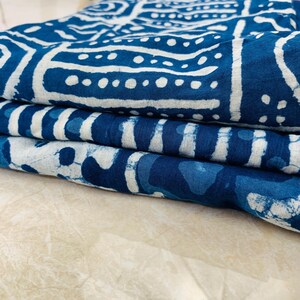 May include: Folded indigo blue fabric with white geometric patterns. The fabric is stacked, showcasing the detailed designs and textures. The patterns include lines, dots, and shapes, creating a visually appealing textile.