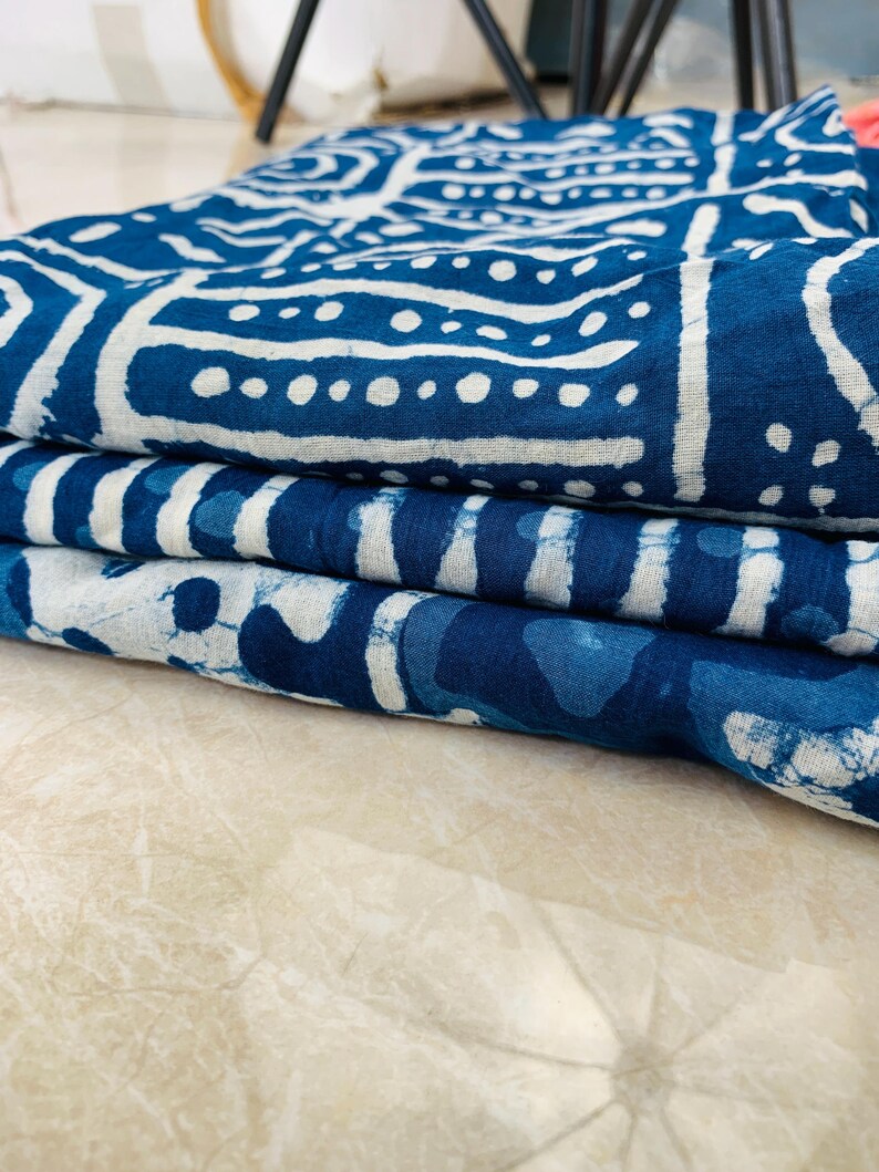 May include: Folded indigo blue fabric with white geometric patterns. The fabric is stacked, showcasing the detailed designs and textures. The patterns include lines, dots, and shapes, creating a visually appealing textile.