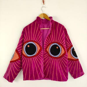 May include: A magenta velvet jacket with embroidered eye designs. The jacket features large, stylized eyes on the front and sleeves, with radiating lines. The eyes have black pupils, blue irises, and red and orange accents. The jacket is hanging on a wooden hanger.