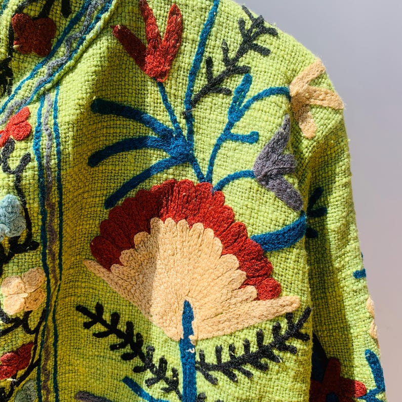 May include: Close-up of a lime green jacket with intricate floral embroidery. The embroidery features red, blue, beige, and black thread, creating a textured and colorful design. The jacket's fabric has a woven appearance.