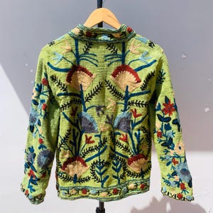 May include: A lime green jacket with long sleeves and a collared neckline. The jacket is adorned with colorful floral embroidery in shades of red, blue, tan, and black. The embroidery covers the front, back, and sleeves of the jacket.