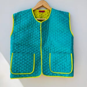 Puede incluir: Un chaleco acolchado turquesa con un patrón de diamantes, ribete amarillo y dos bolsillos delanteros. El chaleco tiene un forro amarillo y cuelga de una percha de madera. Una prenda de abrigo elegante y colorida.