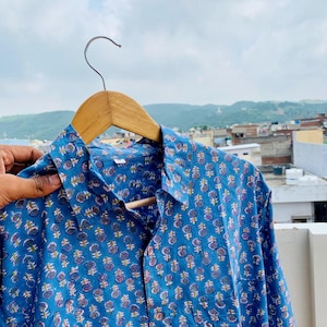 Könnte beinhalten: Ein blaues Hemd mit Knöpfen und floralem Muster, das an einem Holzbügel hängt. Das Hemd hat ein sich wiederholendes Design aus lila und gelben Blumen. Der Hintergrund zeigt eine Stadtlandschaft und einen bewölkten Himmel.