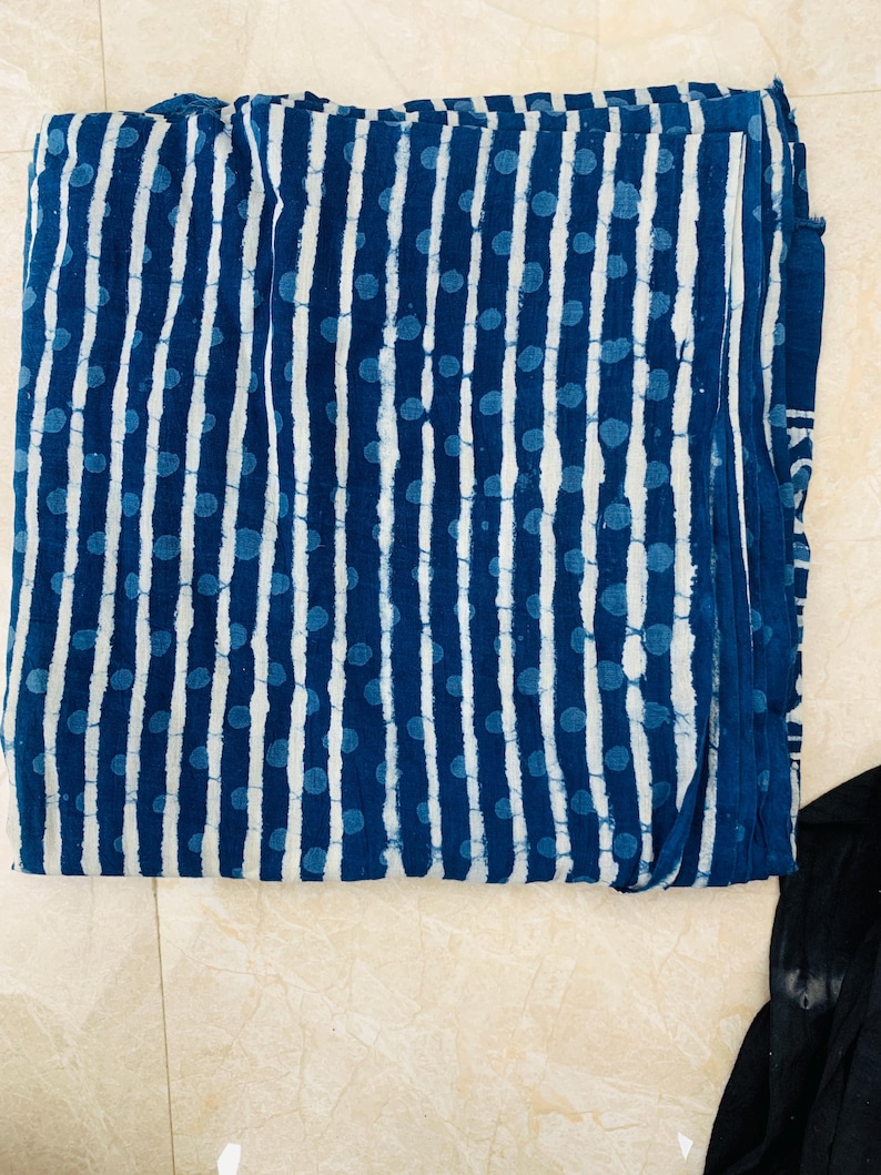 May include: Folded indigo blue fabric with a white vertical stripe and blue dot pattern. The fabric appears to be cotton or a similar natural fiber. The design is reminiscent of traditional block printing.