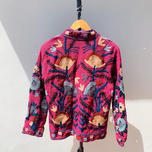 Op de afbeelding: Een magenta jasje met gedetailleerde bloemenborduurwerk. De jas heeft een hoge kraag en lange mouwen, versierd met patronen in blauw, beige en grijs. Het borduurwerk bedekt de rug en de mouwen.