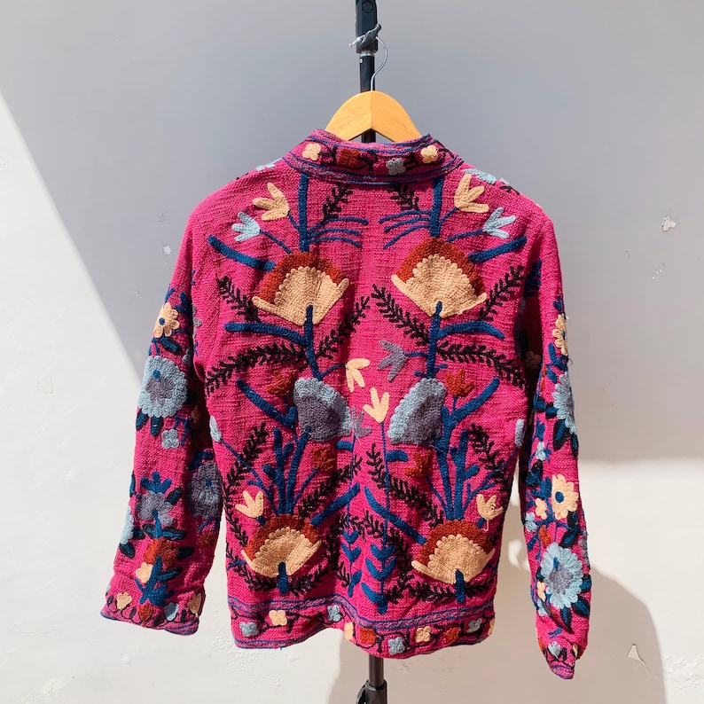 Op de afbeelding: Een magenta jasje met gedetailleerde bloemenborduurwerk. De jas heeft een hoge kraag en lange mouwen, versierd met patronen in blauw, beige en grijs. Het borduurwerk bedekt de rug en de mouwen.
