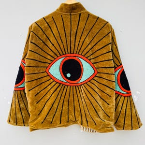 May include: A brown velvet jacket with a large eye design on the back. The eye is outlined in black with a red border and a turquoise iris. The jacket has long sleeves and a collar.