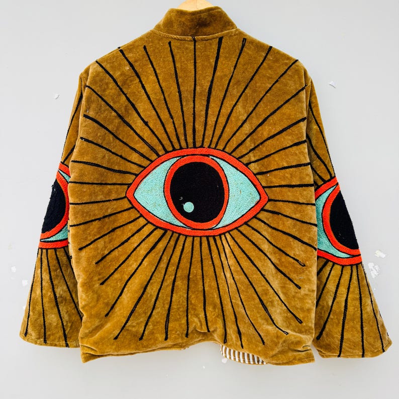 May include: A brown velvet jacket with a large eye design on the back. The eye is outlined in black with a red border and a turquoise iris. The jacket has long sleeves and a collar.
