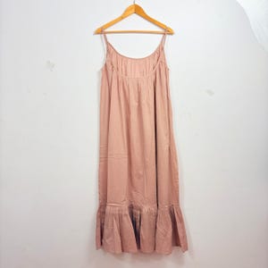 May include: A dusty rose maxi dress with spaghetti straps, hanging on a wooden hanger. The dress features a gathered bodice and a ruffled hemline. The fabric appears to be lightweight and flowing, suitable for warm weather.