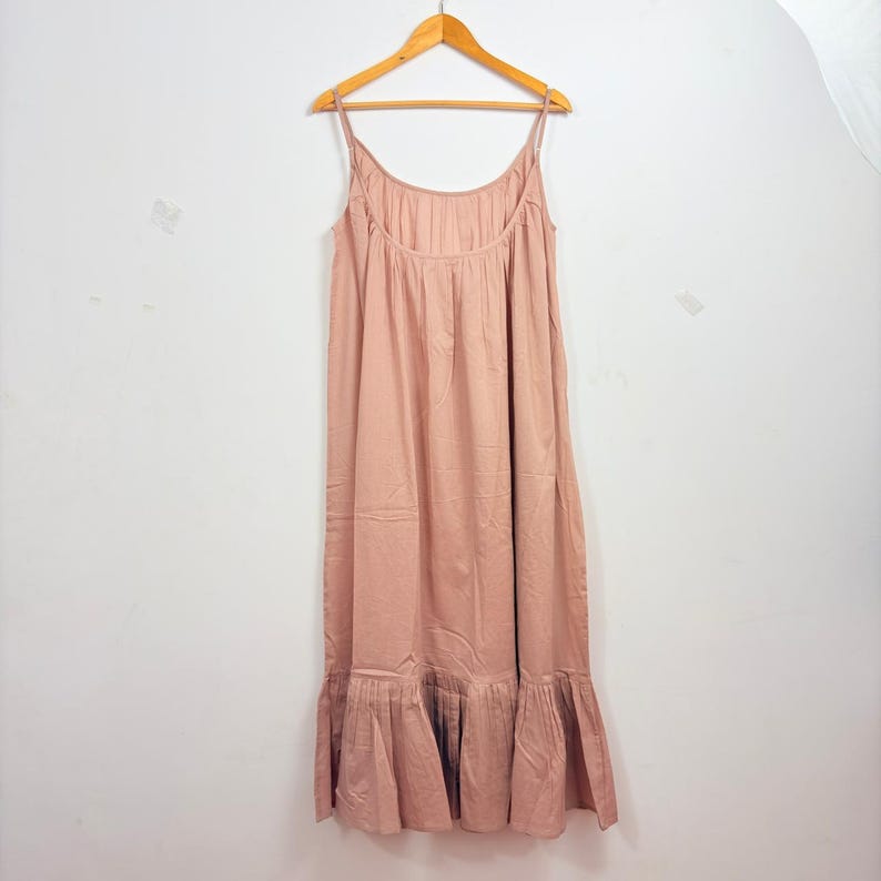 May include: A dusty rose maxi dress with spaghetti straps, hanging on a wooden hanger. The dress features a gathered bodice and a ruffled hemline. The fabric appears to be lightweight and flowing, suitable for warm weather.