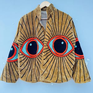 May include: Mustard-colored velvet jacket with large embroidered eye designs in orange, turquoise, and black. The jacket features a high collar and long sleeves, with the eye design repeated on the sleeves. The jacket is hanging on a wooden hanger.