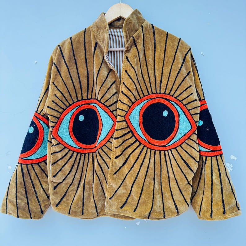 May include: Mustard-colored velvet jacket with large embroidered eye designs in orange, turquoise, and black. The jacket features a high collar and long sleeves, with the eye design repeated on the sleeves. The jacket is hanging on a wooden hanger.