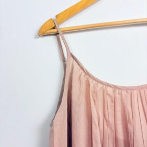 May include: A light pink pleated camisole top with thin spaghetti straps hangs on a wooden hanger against a white wall. The top is made of a soft, flowing fabric and has a simple, elegant design.