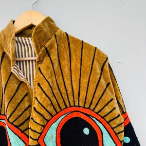 May include: A mustard-colored velvet jacket with black embroidered sunburst designs. The jacket features a high collar and a striped lining. The sleeves have embroidered eye designs with red, black, and turquoise accents. The jacket is hanging on a wooden hanger.