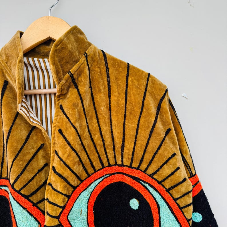 May include: A mustard-colored velvet jacket with black embroidered sunburst designs. The jacket features a high collar and a striped lining. The sleeves have embroidered eye designs with red, black, and turquoise accents. The jacket is hanging on a wooden hanger.