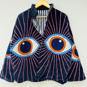 Könnte beinhalten: Marineblaue Samtjacke mit gesticktem Augendesign. Die Jacke hat große, detaillierte Augen in Blau, Orange und Creme mit rosa Strahlen. Das Innenfutter ist schwarz-weiß gestreift.