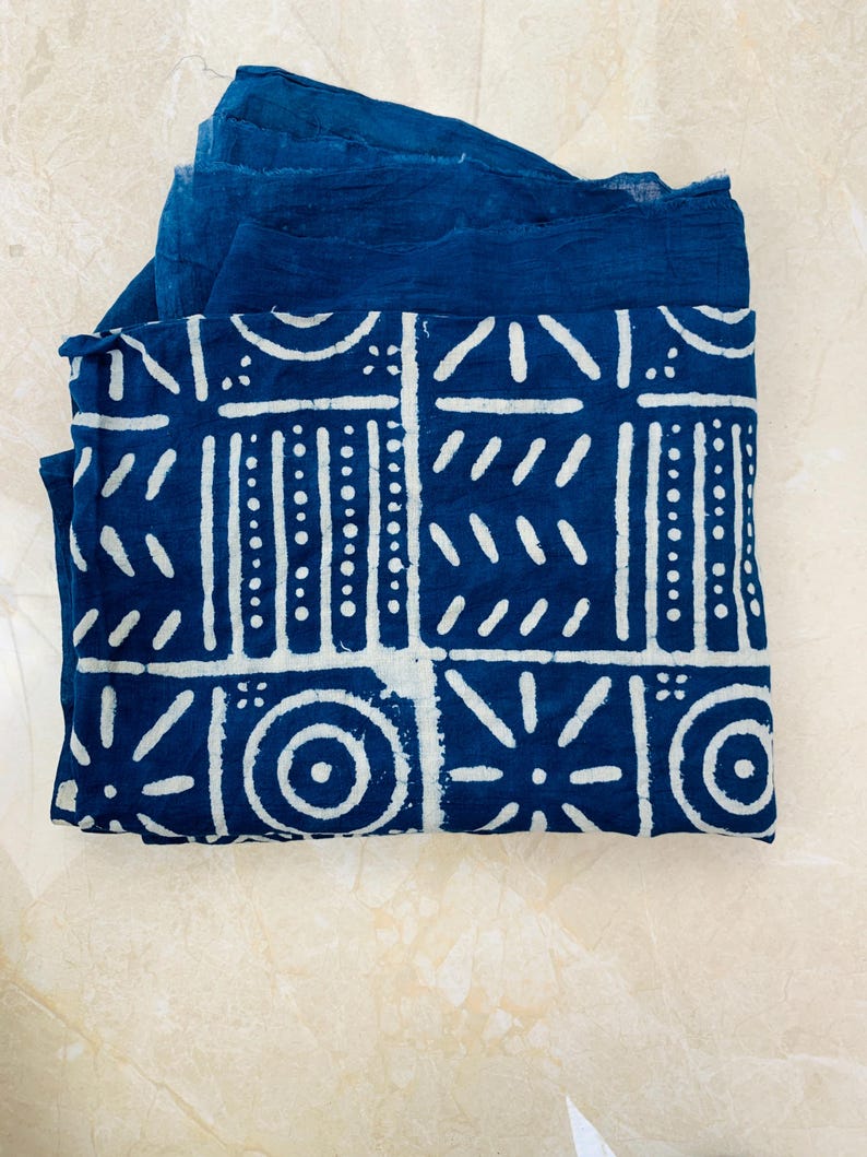 May include: A folded indigo blue fabric with a white geometric pattern. The design features squares, circles, and lines. The fabric is layered with a solid indigo blue textile.