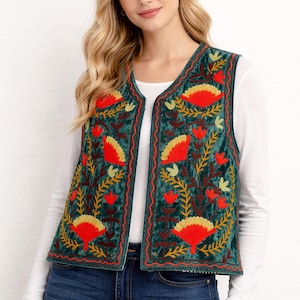 May include: A teal velvet vest with a floral embroidery design in red, yellow, and gold. The vest has a V-neck and is worn over a white long-sleeved shirt and blue jeans. The floral pattern is repeated across the vest.