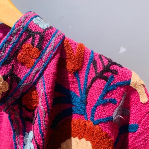 Op de afbeelding: Close-up van een felroze geborduurde jas. De jas heeft ingewikkelde bloempatronen in tinten oranje, blauw en beige. De borduursels zijn gedetailleerd en geven het kledingstuk textuur. De jas wordt op een houten hanger getoond.
