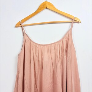 May include: A light pink camisole dress with thin straps hangs on a wooden hanger. The dress has a gathered neckline and a flowing, pleated design. The fabric appears to be a soft, lightweight material.