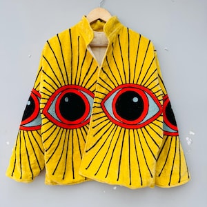 Könnte beinhalten: Eine gelbe Samtjacke mit einem einzigartigen Augendesign. Die Jacke hat große, gestickte Augen mit schwarzen Pupillen, roten Umrissen und blauen Iris. Schwarze Linien strahlen von den Augen aus und erzeugen einen Sonnenstrahleffekt. Die Jacke hängt an einem Holzkleiderbügel.